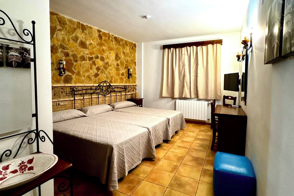 Une chambre avec deux lits, un mur en pierre, un radiateur, une table avec une carafe et un pouf bleu.
