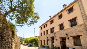 Vista de una calle con un edificio de fachada de piedra, varias ventanas y un área de mesas al aire libre.