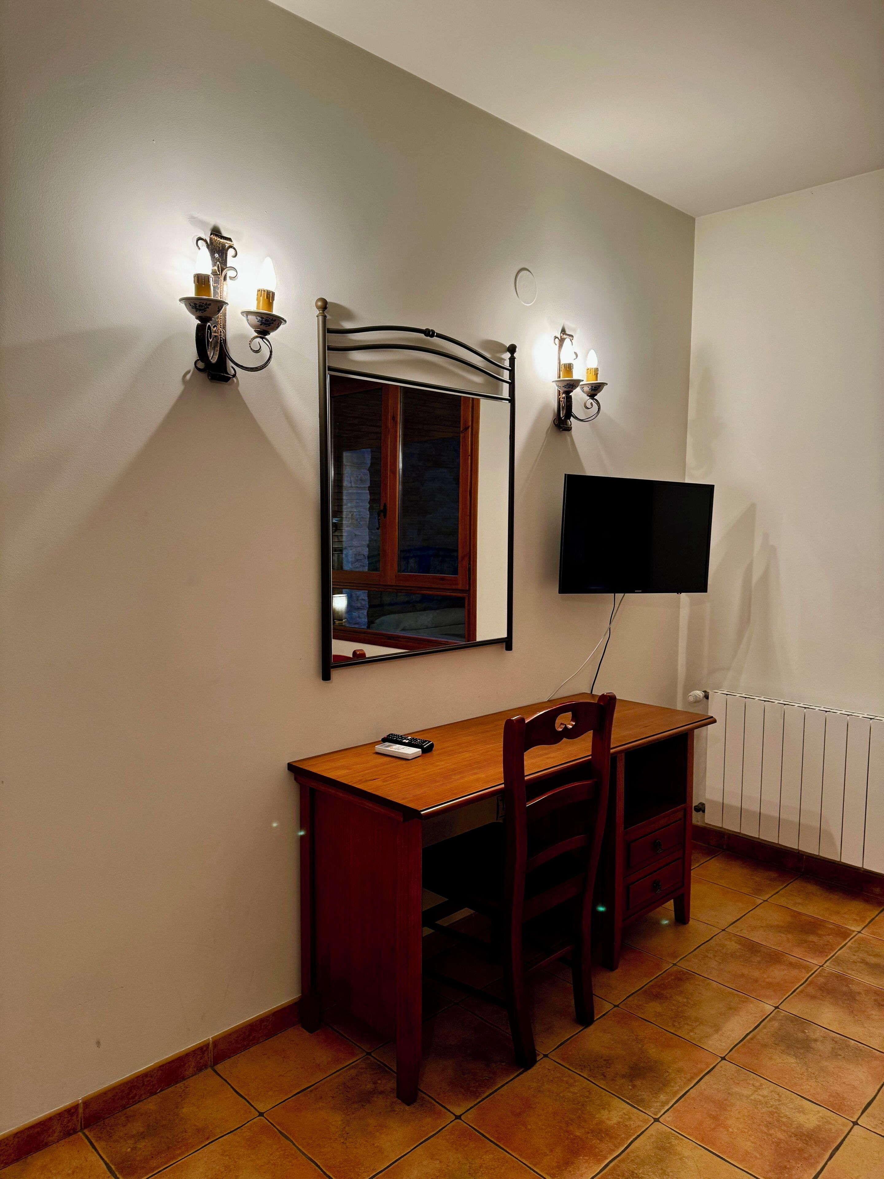A room with a wooden desk, a chair, a television mounted on the wall, and two wall-mounted sconces.