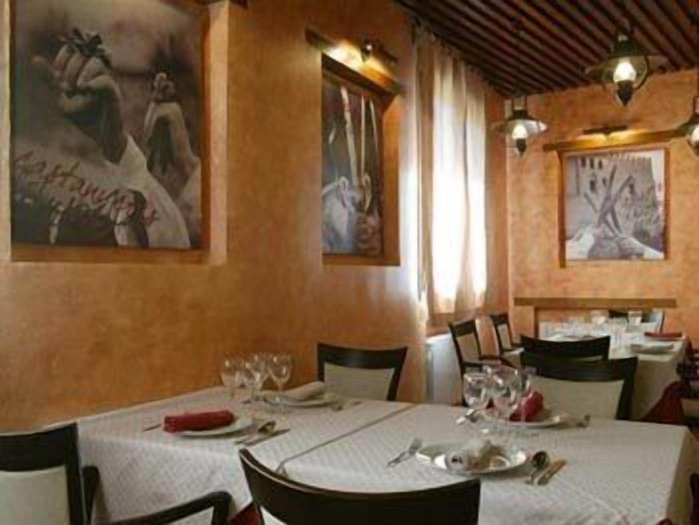 A dining area with round tables set for four, each with white tablecloths, plates, and cutlery. There are framed artworks on the walls, and the room has a warm, wooden ceiling.
