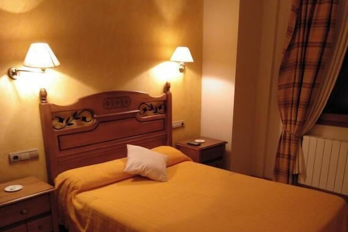 Une chambre avec un lit simple, une tête de lit en bois, deux lampes de chevet, une table de chevet, une petite table et une fenêtre avec des rideaux.