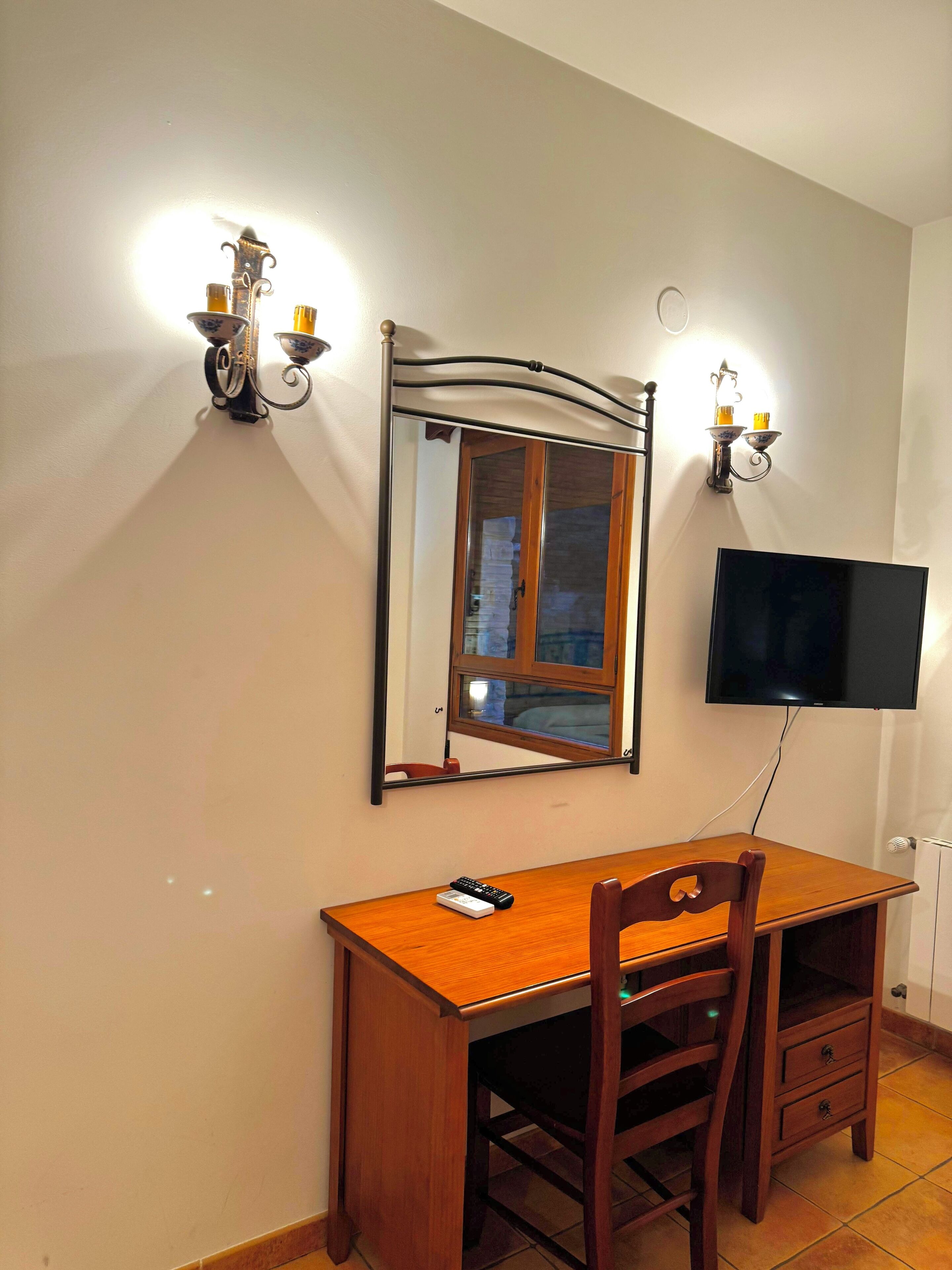 Una habitación con un escritorio de madera, una silla, un televisor de pantalla plana montado en la pared y dos apliques de pared con velas.