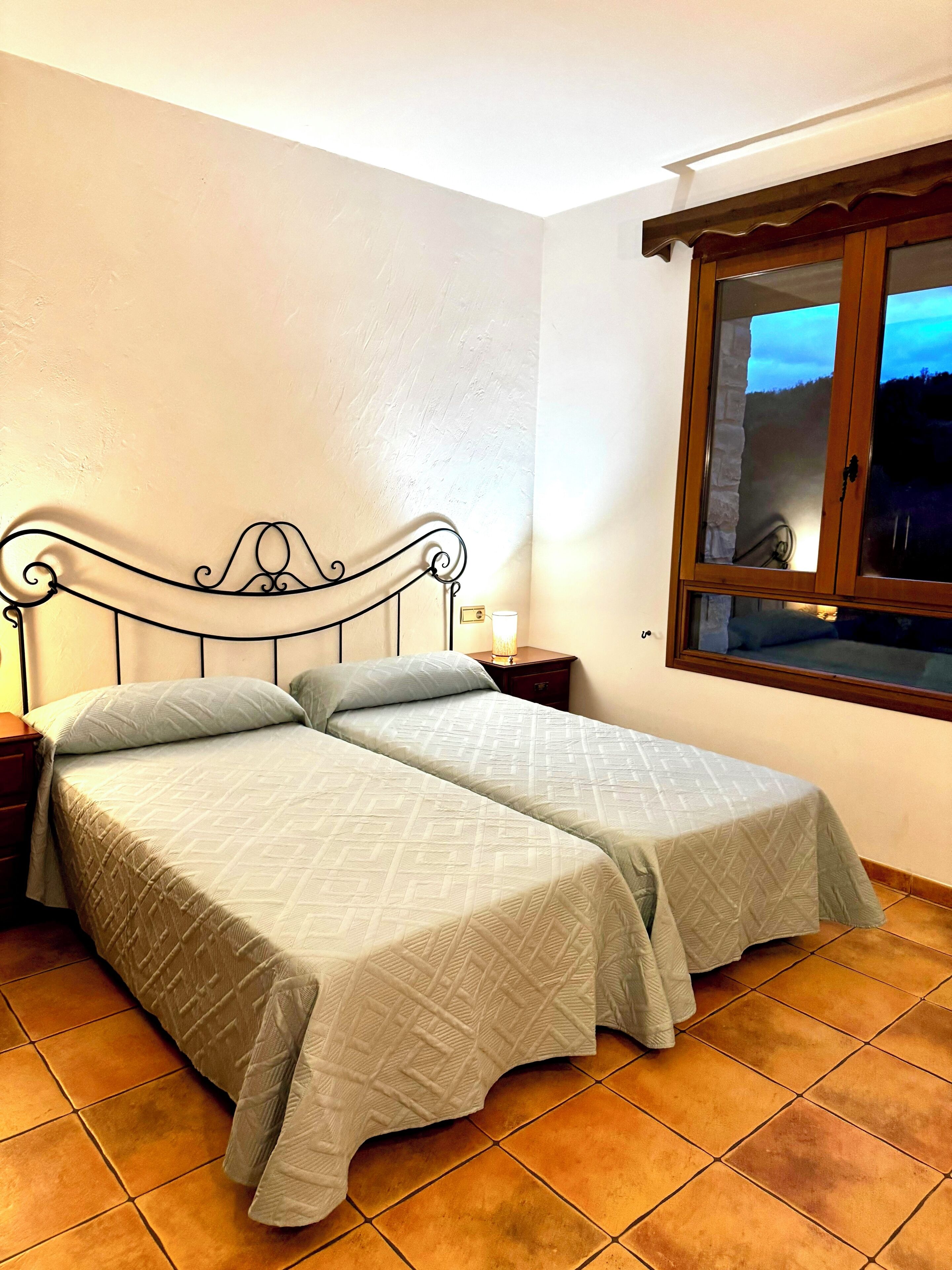 A double bed with a metal frame and two pillows, a bedside table with a lamp, and a window with a view of the sky.