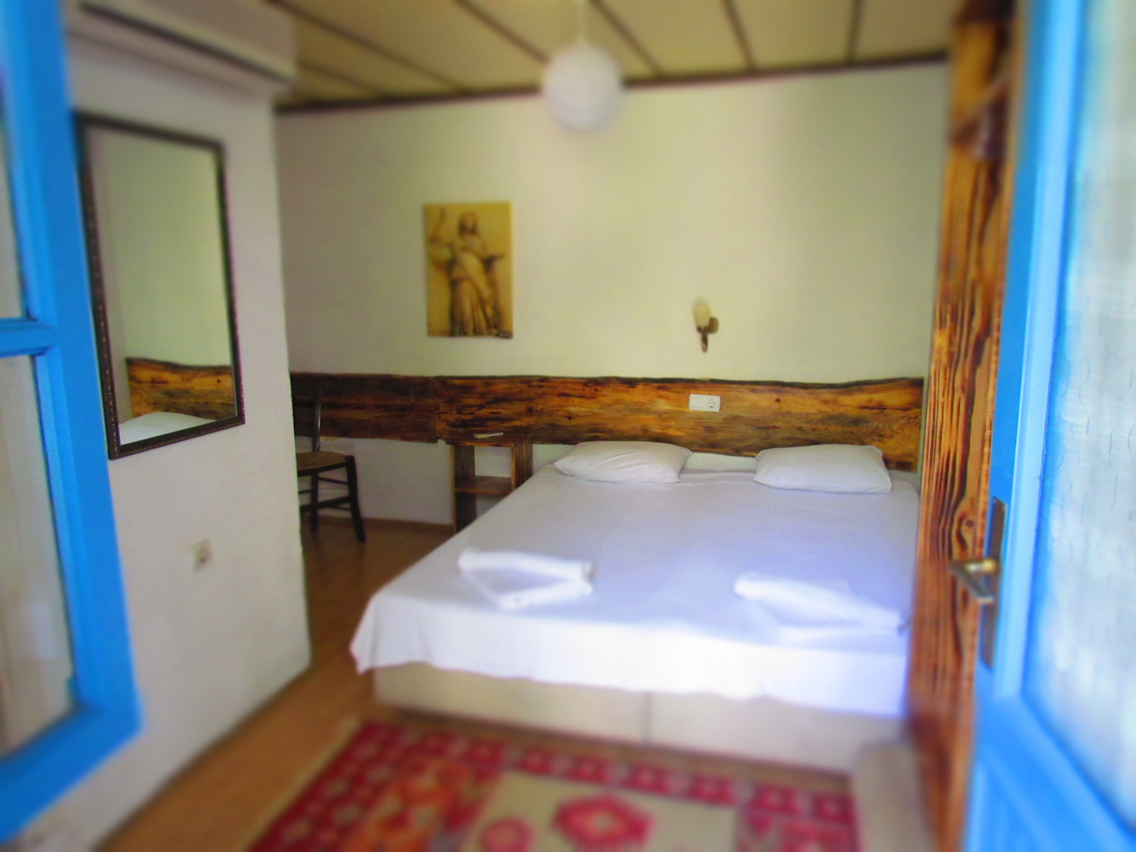 A bedroom with a wooden headboard, a bed with white linens, a chair, a wall-mounted light, and a painting.