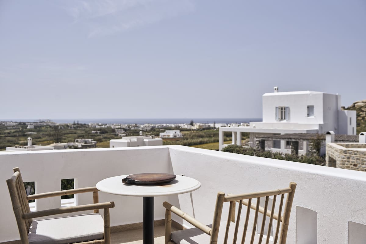 A balcony with a table and two chairs, overlooking a coastal town with white buildings and a clear sky.
