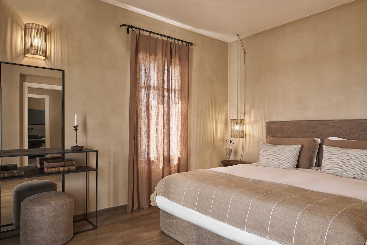 A hotel room with a large bed, a bedside table with a lamp, a mirror, and a desk with books.