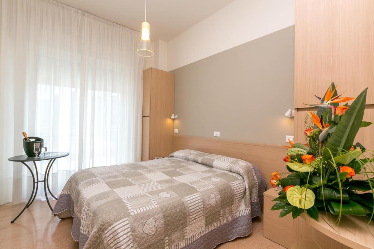 A hotel room with a bed, a small table with a drink, a decorative plant, and a wooden cabinet.