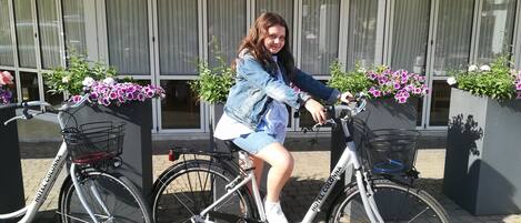 Una persona montando en bicicleta con una cesta delantera frente a un edificio con ventanas amplias y macetas con flores.