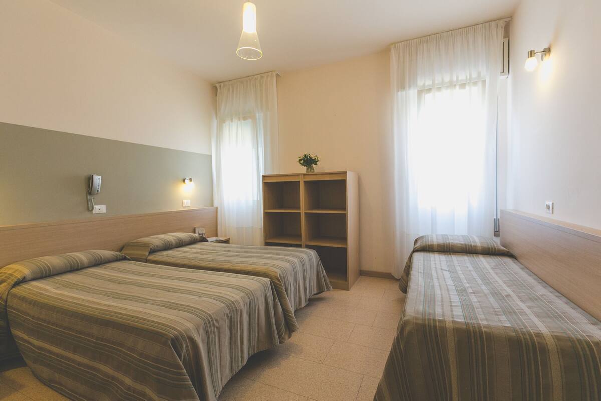 A hotel room with two single beds, a small desk, a shelf unit, and a window with curtains.