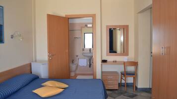 Habitación de hotel con cama, escritorio, silla, armario y baño visible a través de una puerta abierta.