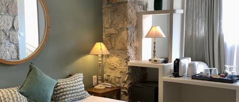 Una habitación de hotel con una cama, una mesita de noche, una lámpara, un espejo, una pared de piedra y un estante con artículos de aseo.