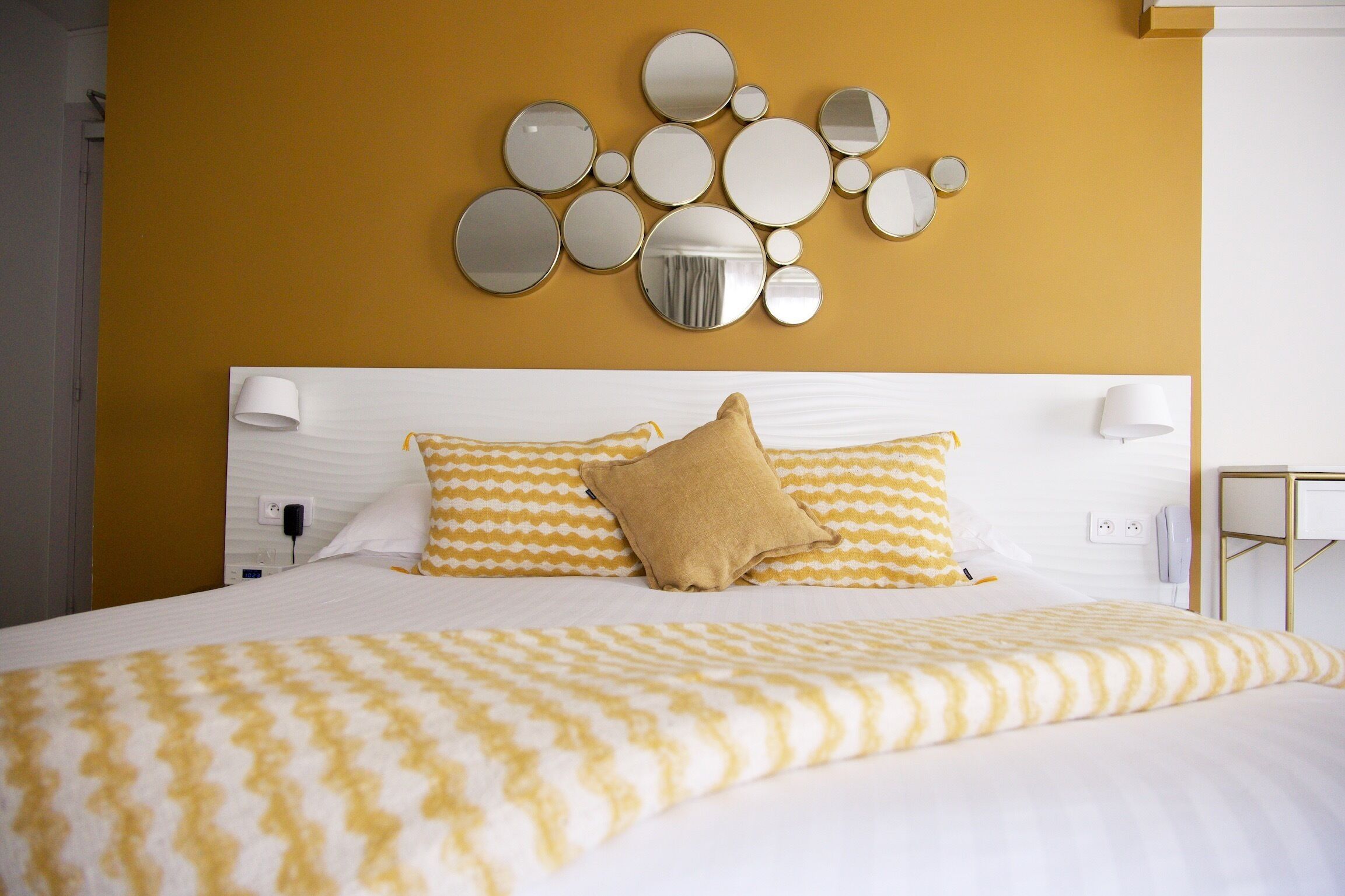 A hotel room with a bed, white headboard, yellow and white patterned bedding, a wall-mounted mirror, and a bedside lamp.