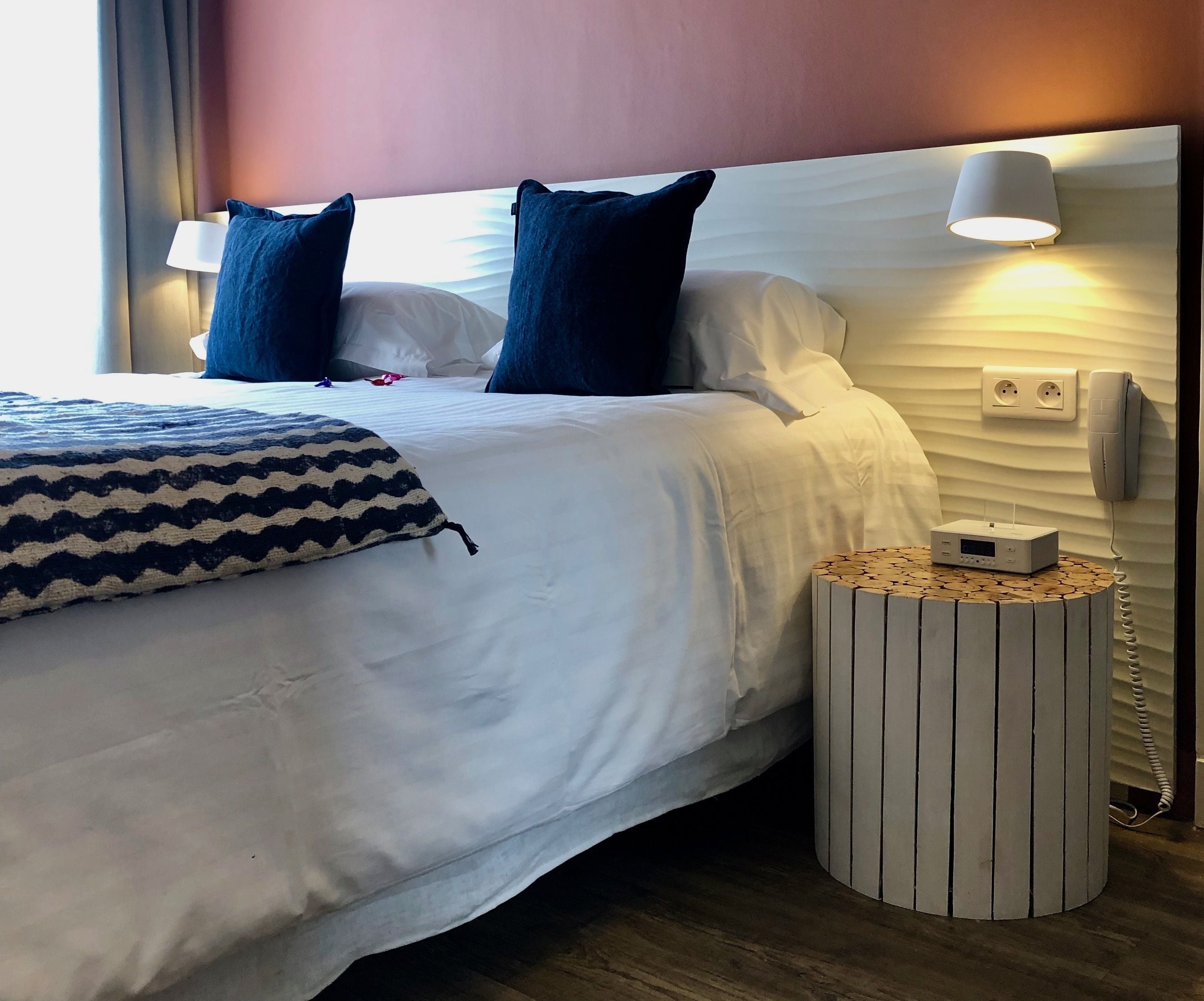A neatly made bed with white bedding and two dark blue pillows, a bedside lamp, a small wooden side table with a white device, and a wall-mounted phone.