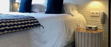 A neatly made bed with white bedding and two dark blue pillows, a bedside lamp, a small wooden side table with a white device, and a wall-mounted phone.