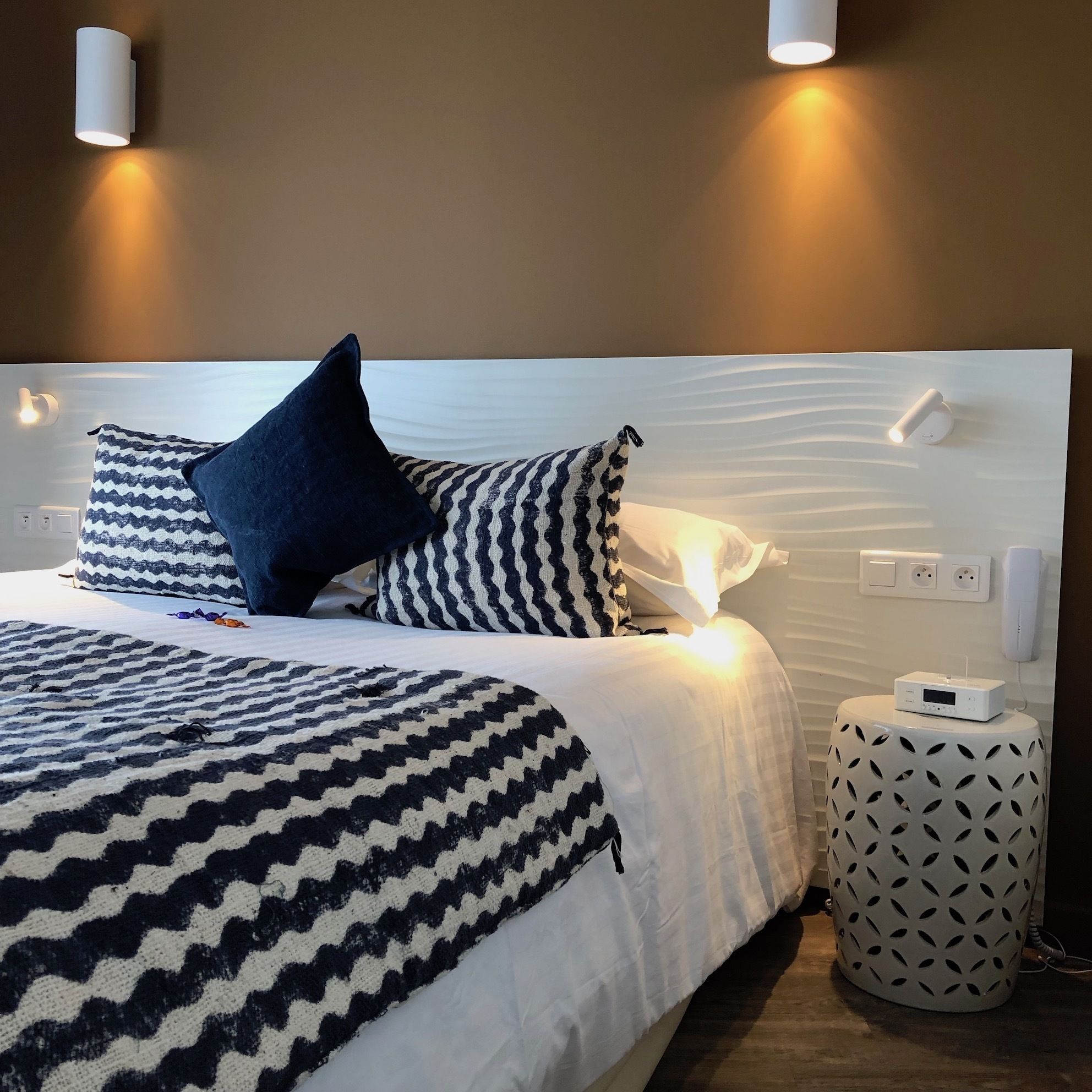 A neatly made bed with patterned bedding, a bedside table with a radio, and two wall-mounted lights.