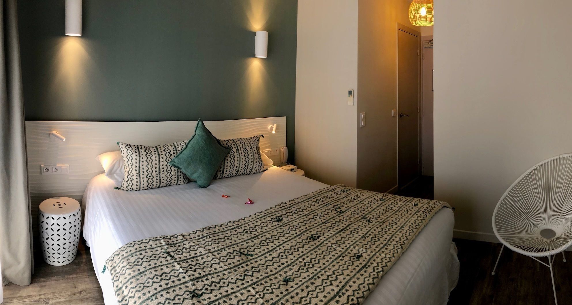 A neatly made bed with patterned bedding, two wall-mounted lights, a bedside table with a patterned lamp, and a chair with a woven seat.