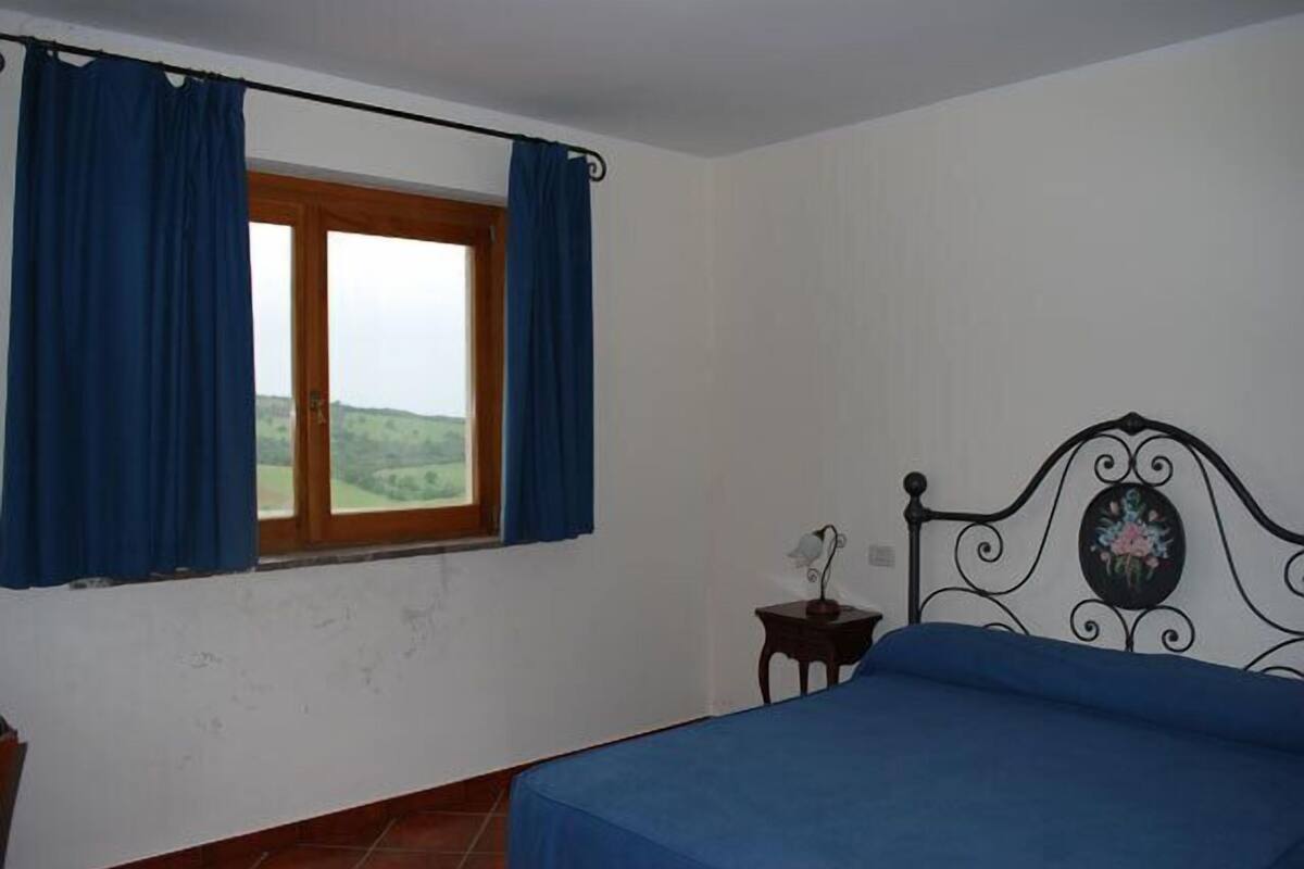 Ein Schlafzimmer mit Bett, Vorhängen am Fenster, einem kleinen Nachttisch mit Lampe und einem dekorativen Kopfteil.