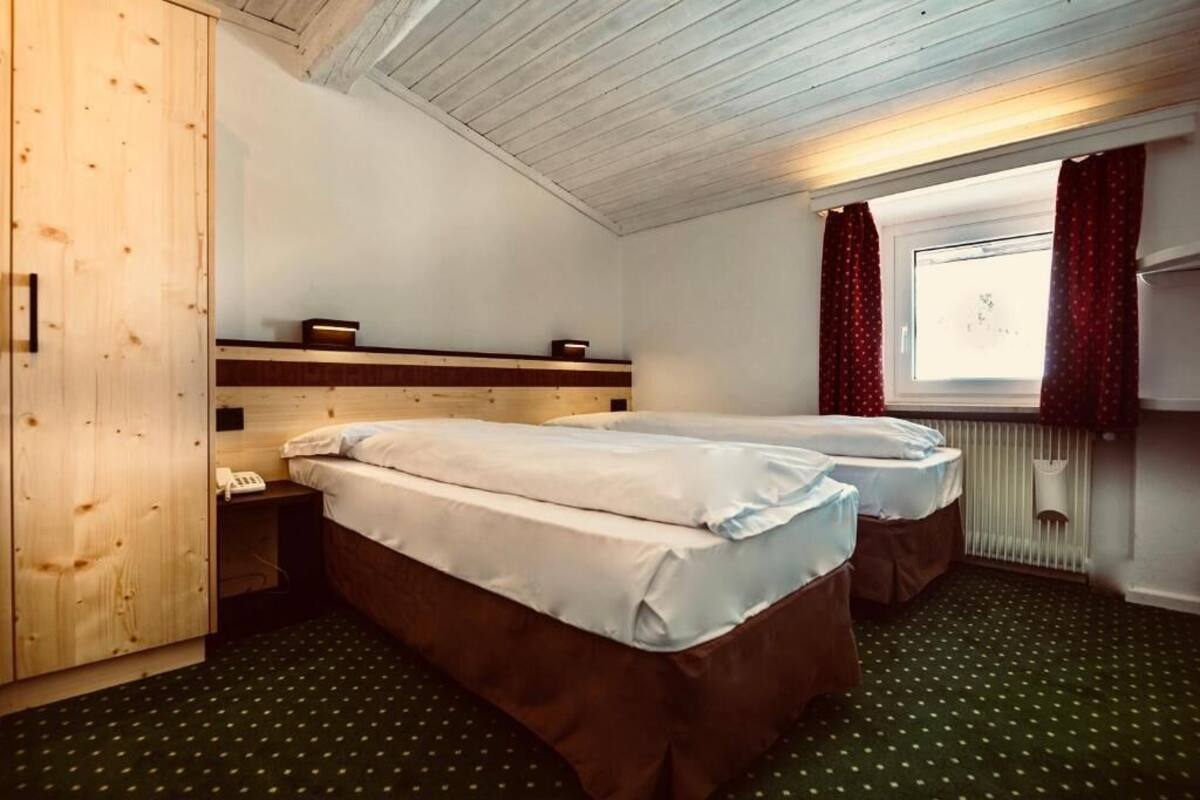 Une chambre avec deux lits simples, une armoire en bois, une petite table de chevet avec un téléphone et une fenêtre avec des rideaux.