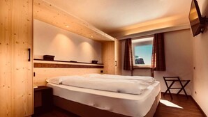 A modern hotel room with a wooden floor, a large bed with white linens, a small bedside table, a window with curtains, and a mounted TV. - Hotel Cir (Corvara in Badia)