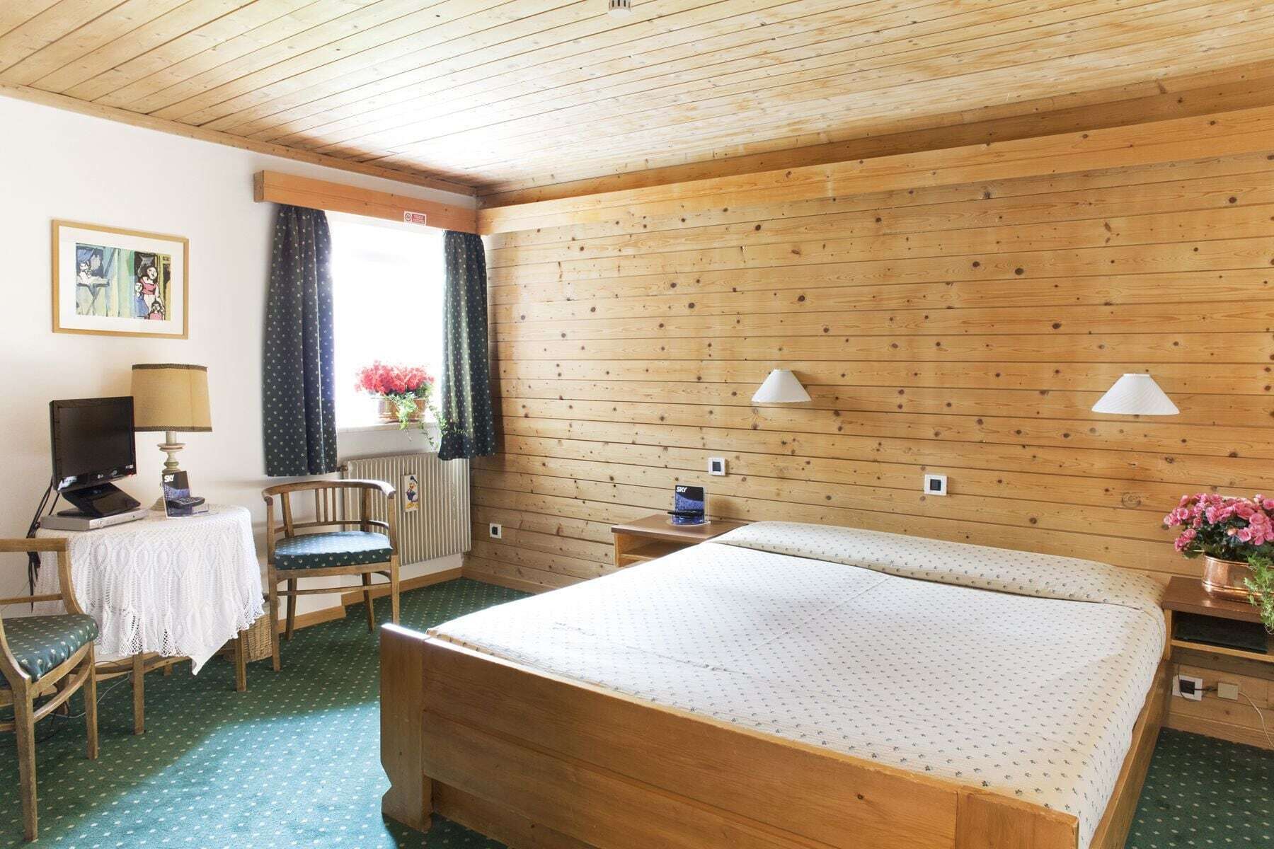 A cozy room with a wooden bed, a desk with a lamp, a chair, and a window with curtains.