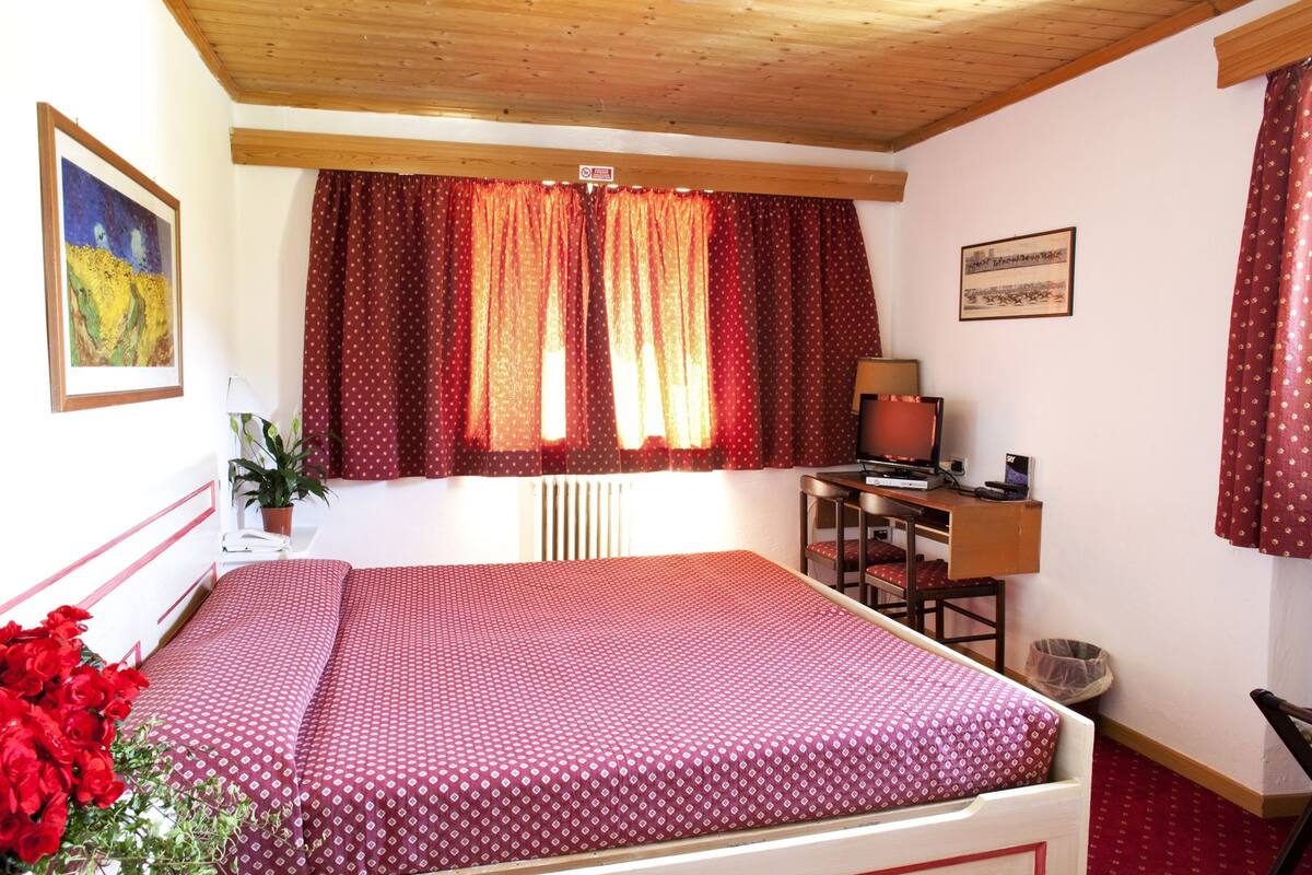 Une pièce comprenant un lit, un bureau avec un ordinateur, une chaise, un radiateur et une fenêtre avec des rideaux.