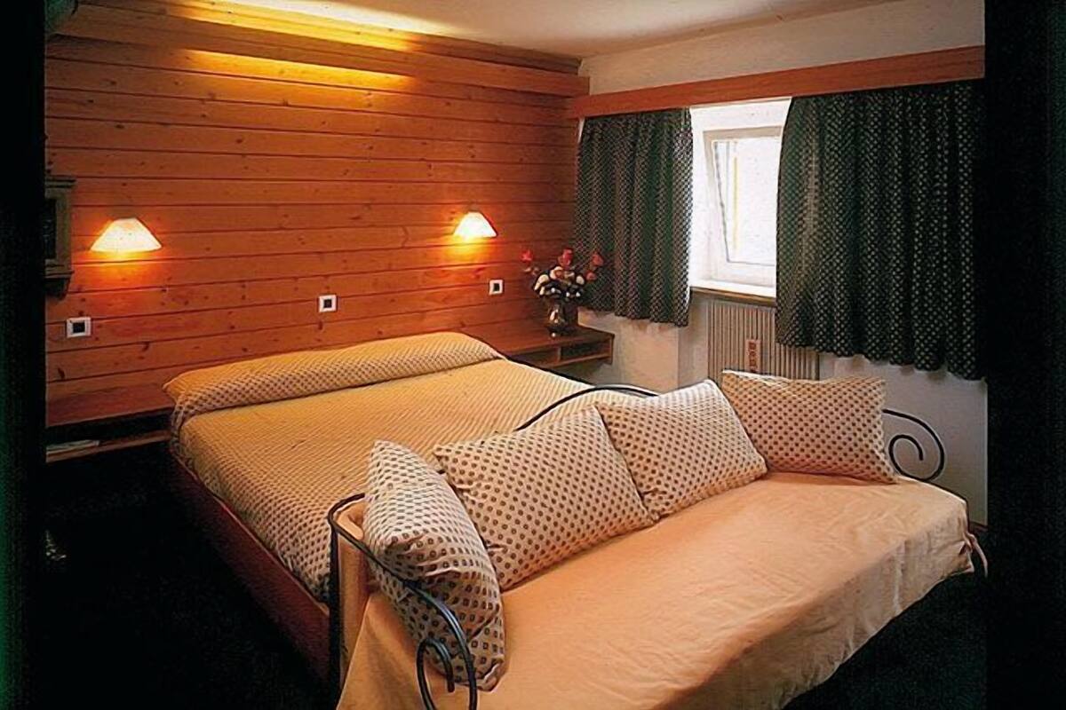Une pièce de type cabane en bois, avec un lit, deux lampes fixées au mur et une fenêtre avec des rideaux.