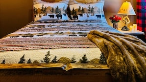 Una cama con edredón estampado, cabecera de madera, lámpara de noche y un florero con flores rojas.