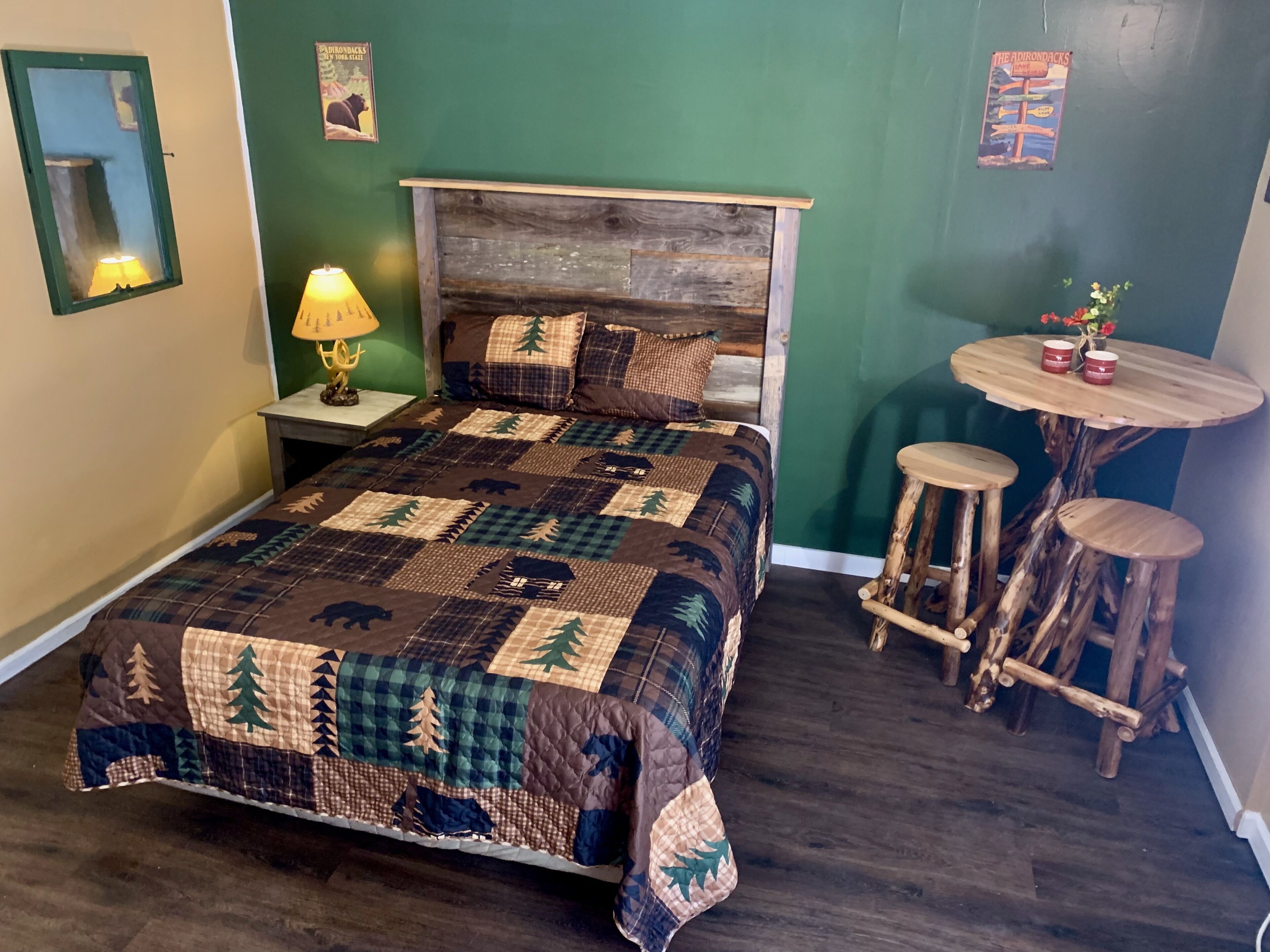 Un dormitorio con una cama grande, una mesita de noche con lámpara, un cabecero de madera, una pequeña mesa redonda con taburetes y una pared verde con carteles.