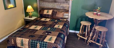 Un dormitorio con una cama grande, una mesita de noche con lámpara, un cabecero de madera, una pequeña mesa redonda con taburetes y una pared verde con carteles.