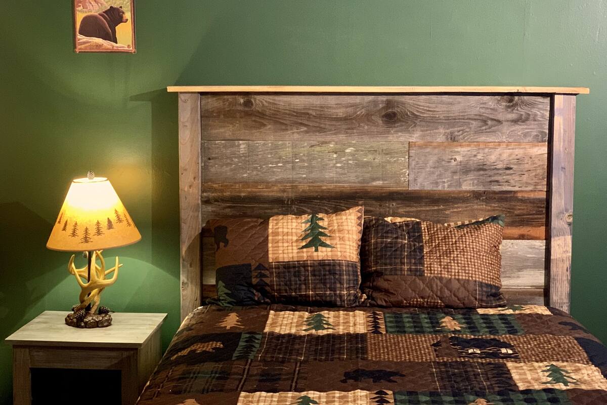 Un dormitorio con cabecera de madera, colcha acolchada con motivos de bosque, una lámpara sobre una mesita de noche y un cartel en la pared.
