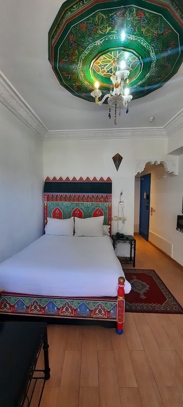 A bedroom with a large bed, a chandelier, a decorative ceiling, and a small table with a lamp.