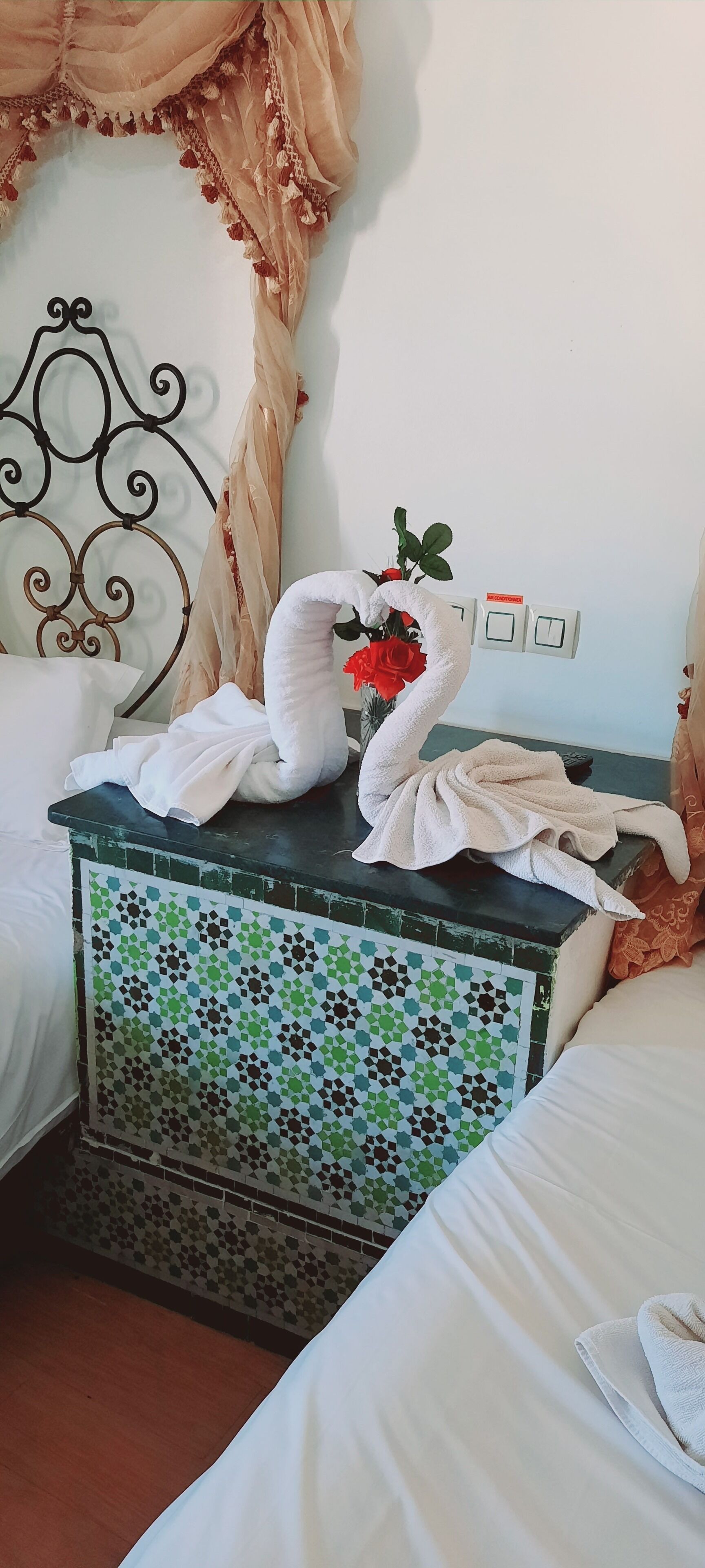 A neatly made bed with white linens, a decorative metal headboard, a bedside table with a mosaic tile design, and a wall-mounted light switch.