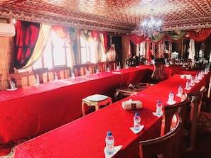 Sala de conferências com mesas compridas cobertas por tecido vermelho, equipada com microfones e garrafas de água.