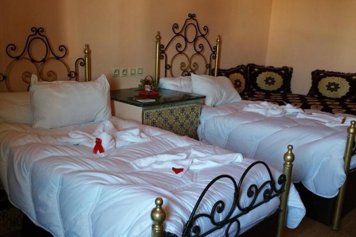 Ein Doppelbett mit Metallgestell und weißer Bettwäsche, ein kleiner Nachttisch mit einer Tablettablage mit grünem und goldenem Muster und eine Wand mit floralem Dekor.