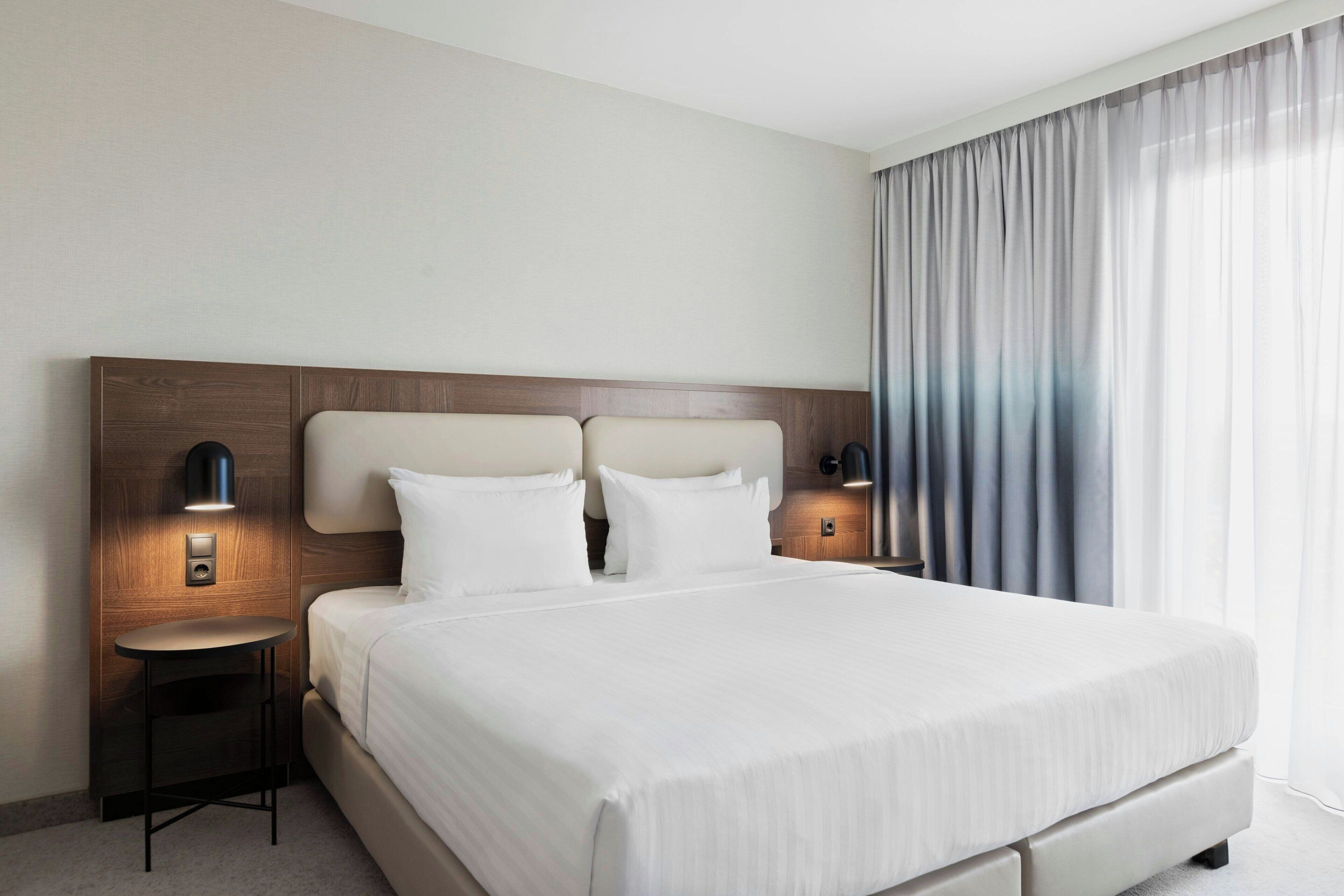 A modern hotel room with a large bed, two bedside tables with lamps, a wooden headboard, and a view of the outdoors through sheer curtains.