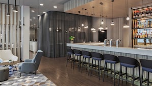 A modern bar area with a bar counter, multiple hanging pendant lights, and a selection of bottles on shelves. - Courtyard München City Ost (Munich)