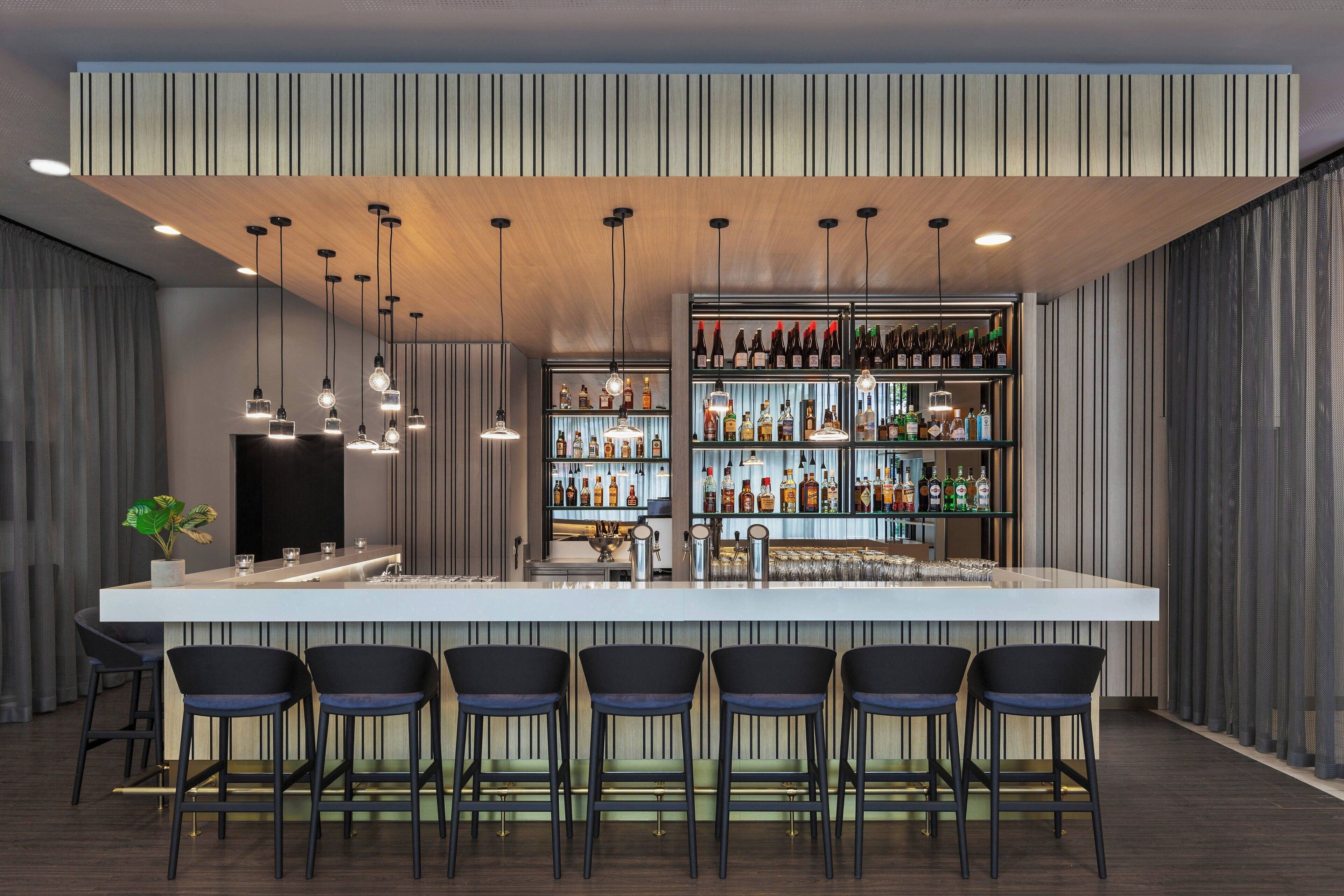 A modern bar area with a long counter, multiple hanging pendant lights, and a variety of bottles and glasses on shelves.