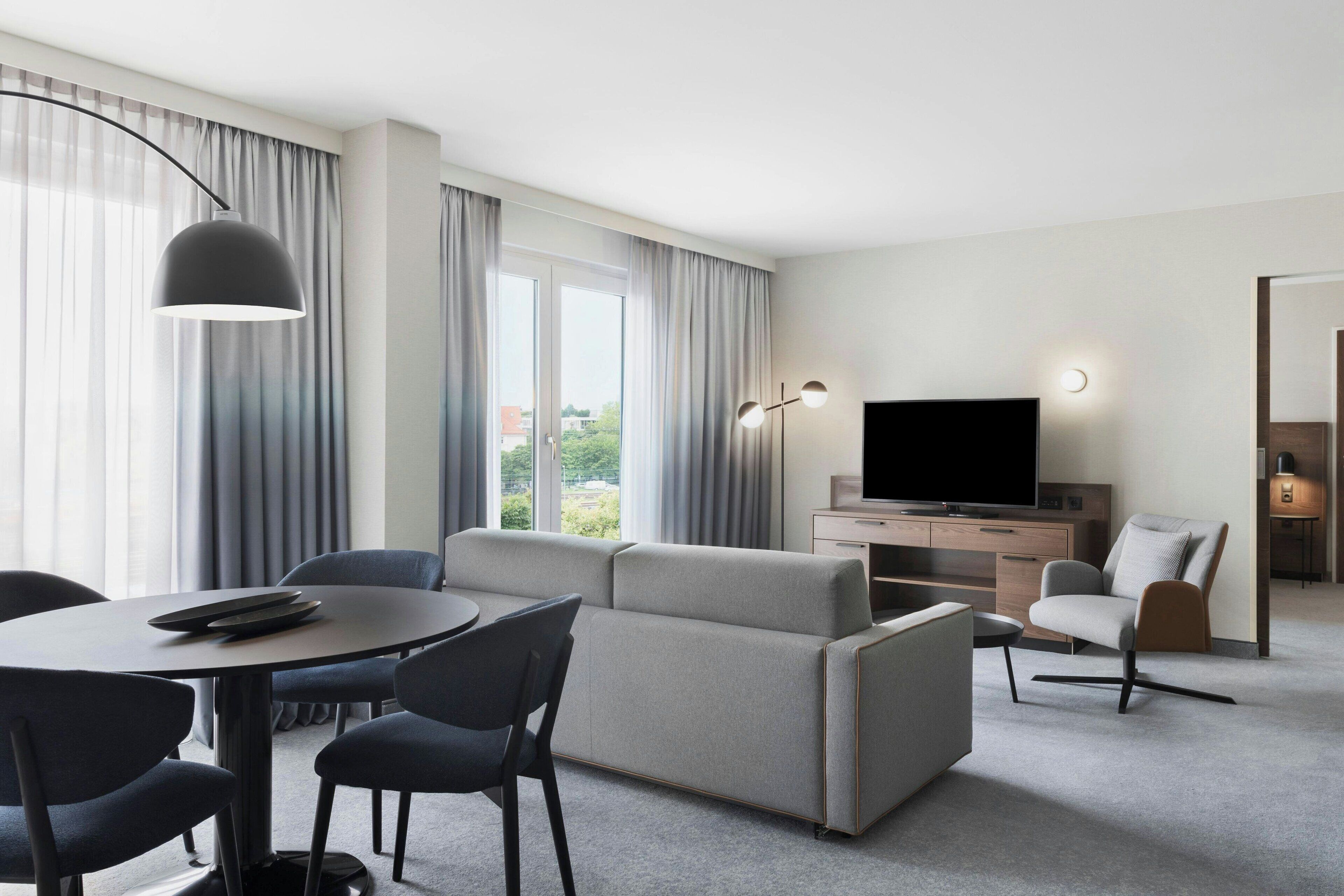 Una habitación de hotel moderna con un sofá gris, una mesa de comedor con sillas, un televisor y un escritorio con una lámpara.