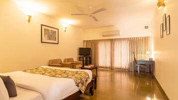 A hotel room with a bed, two chairs, a desk, a television, and a ceiling fan.