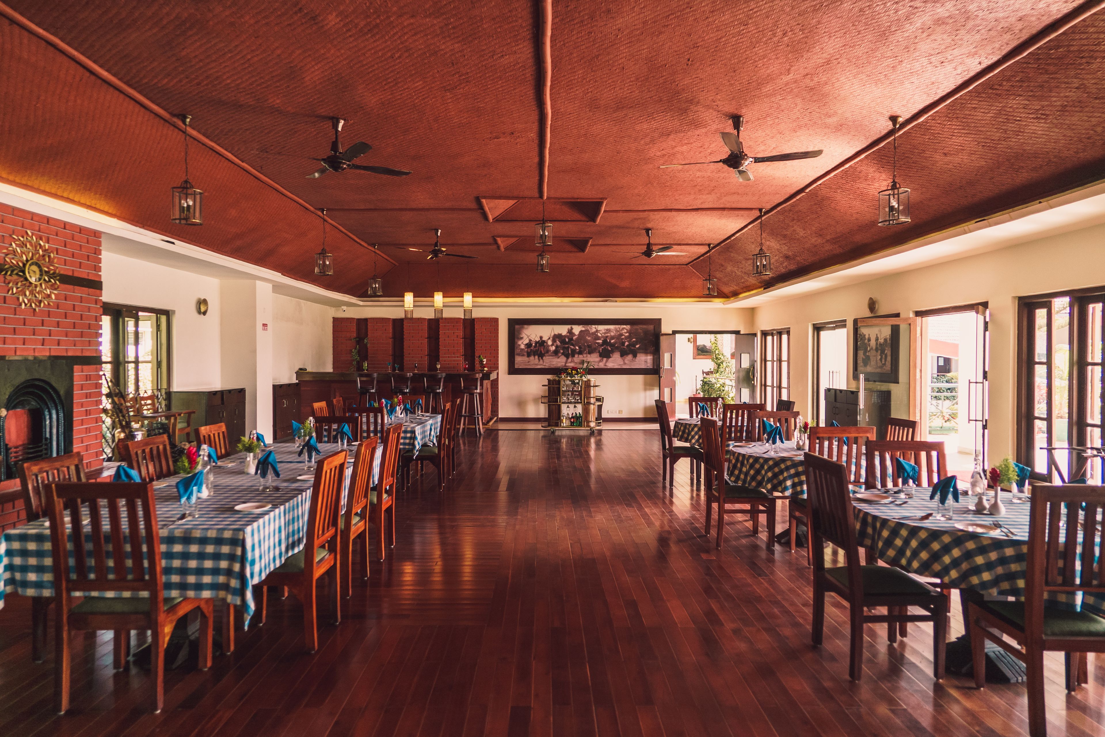 A spacious dining area with wooden floors and tables set for dining, featuring ceiling fans and hanging light fixtures.