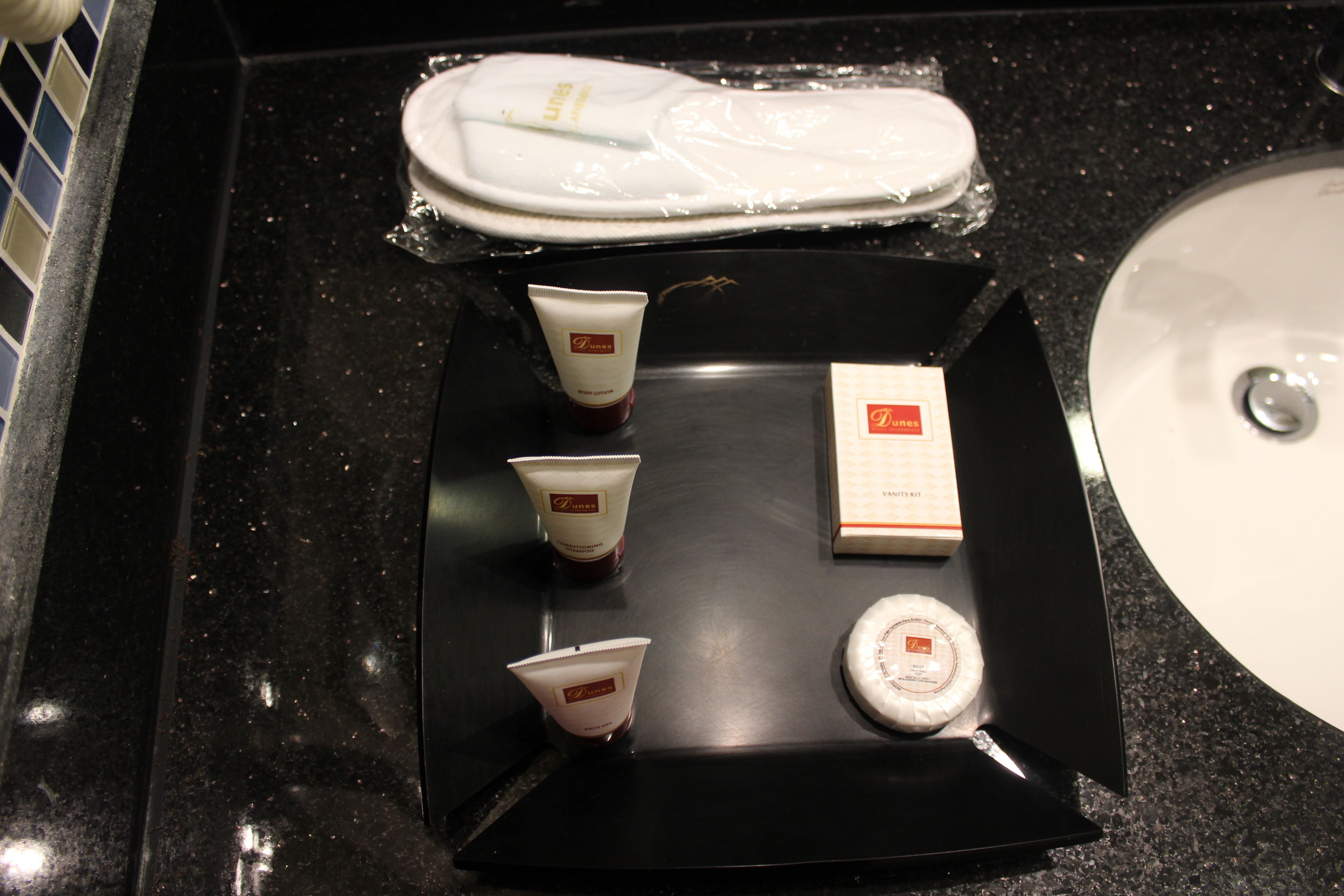 Una bandeja con artículos de aseo personal, incluyendo dos tubos de loción, una pastilla de jabón y un pequeño recipiente, sobre una encimera negra cerca de un lavabo.