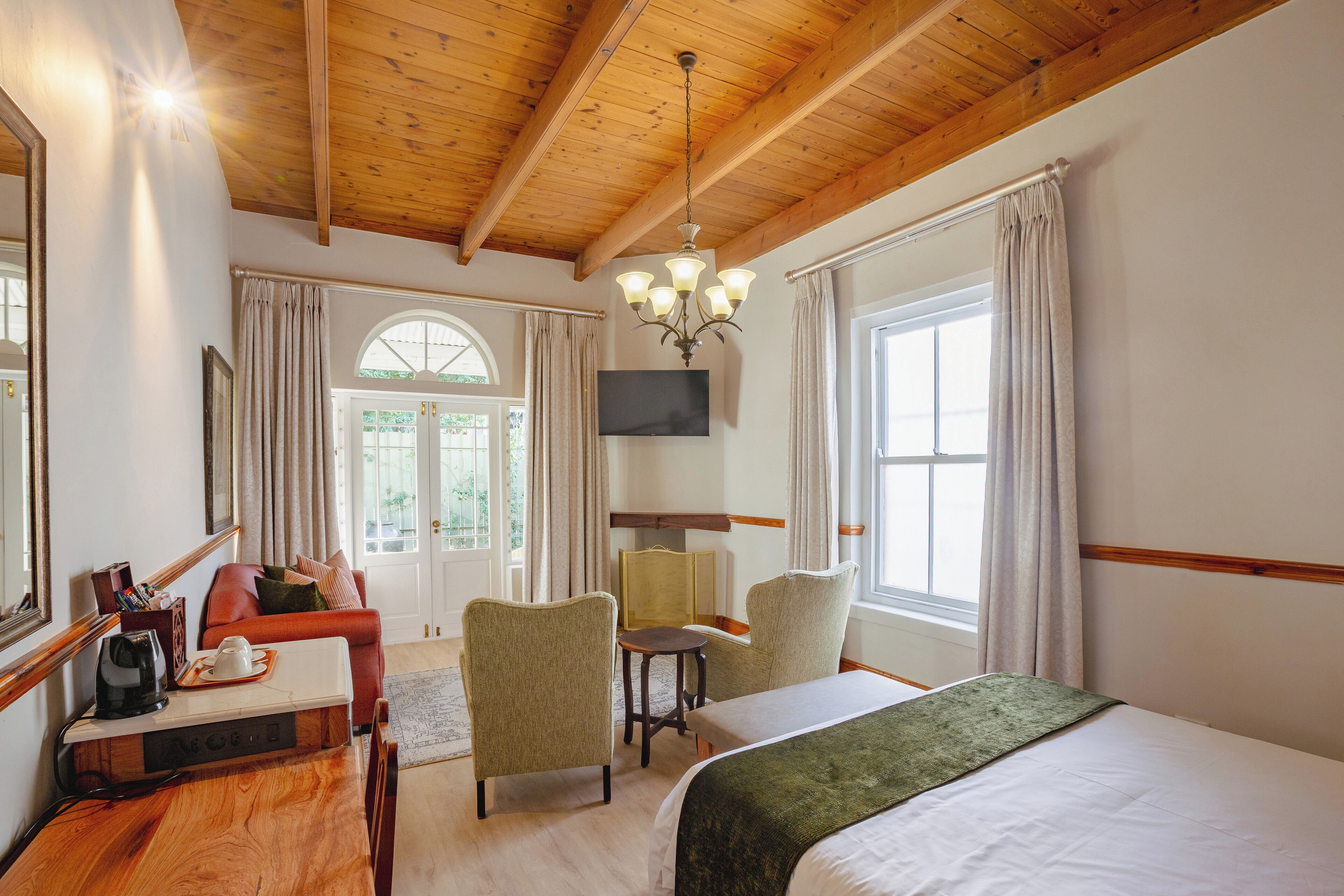 Une chambre d’hôtel avec un plafond en bois, un lit, un bureau avec une bouilloire, deux chaises, une télévision et de grandes fenêtres avec des rideaux.