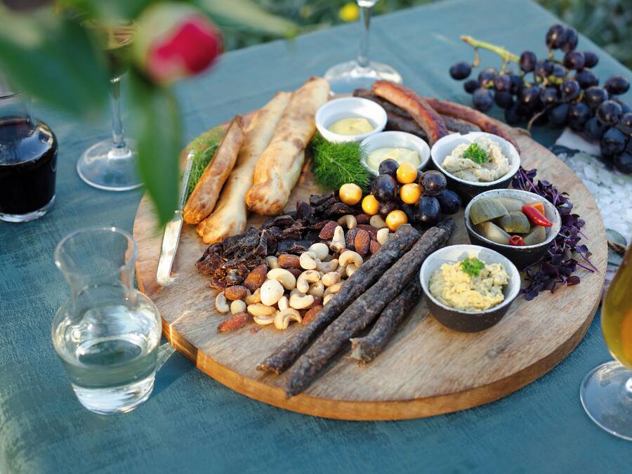 Un tagliere di legno con diversi formaggi, noci e frutta, accompagnato da bicchieri di vino e una bottiglia d'acqua.