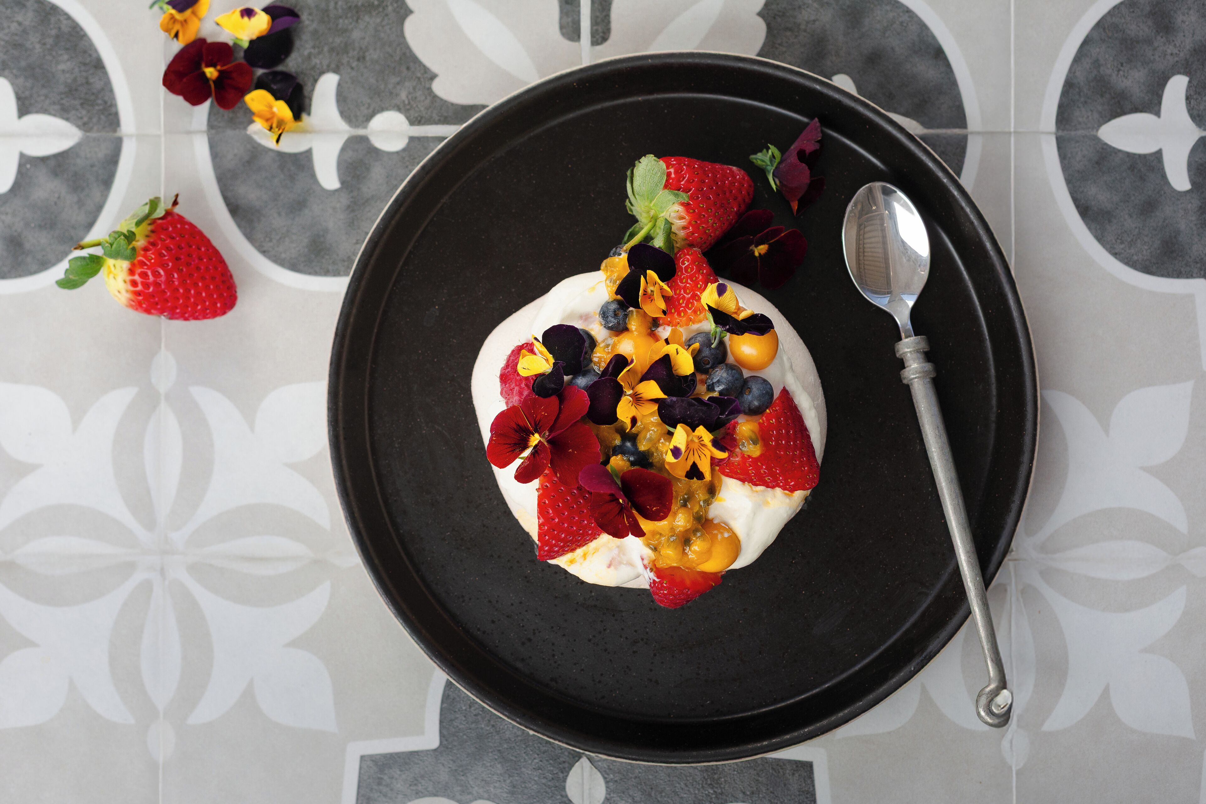 A dessert on a black plate with strawberries, blueberries, and edible flowers, accompanied by a spoon.