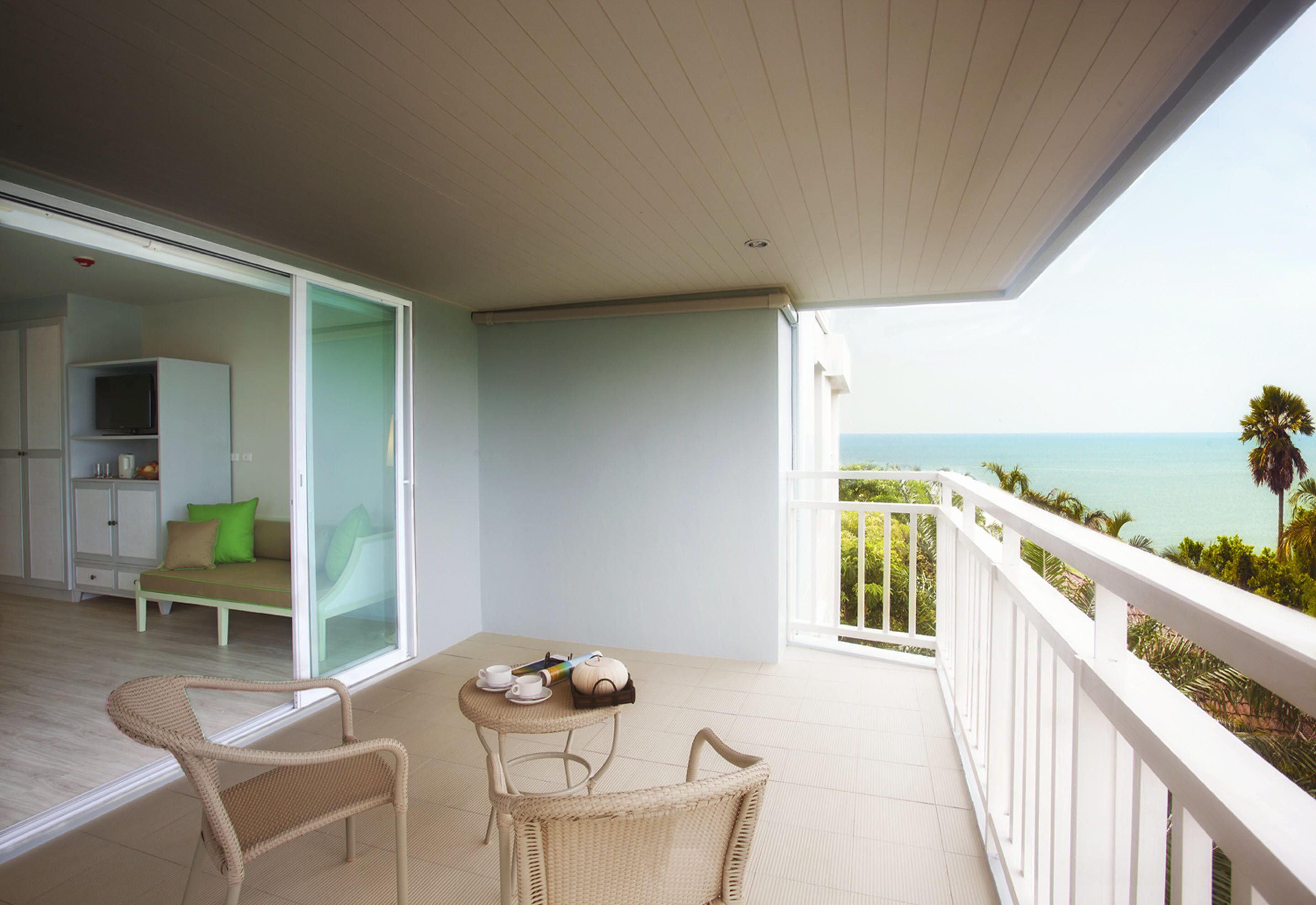 Uma varanda com vista para o mar, um sofá, uma mesa com chávenas e um bule, e cadeiras de vime.