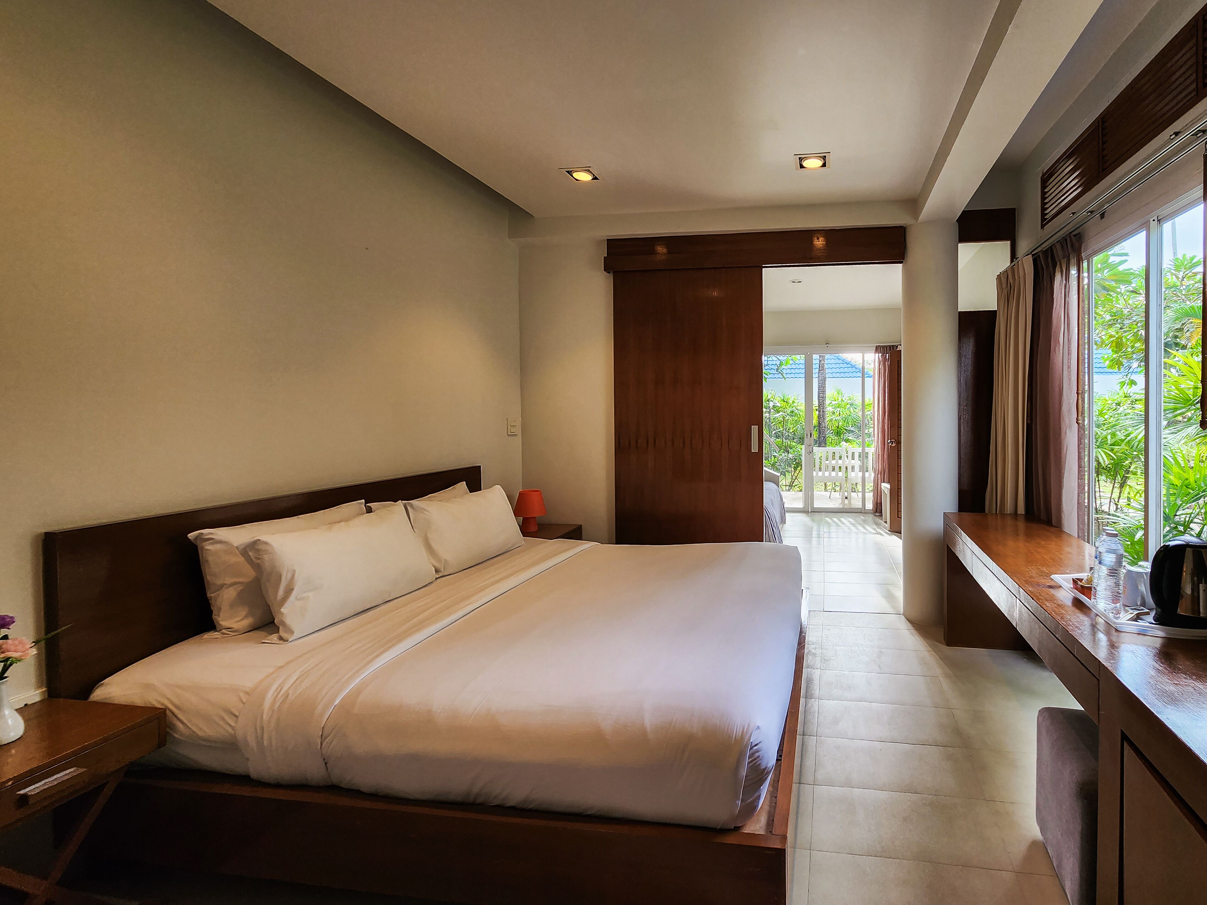 A hotel room with a large bed, a wooden headboard, a nightstand with a lamp, a window with curtains, and a view of greenery outside.