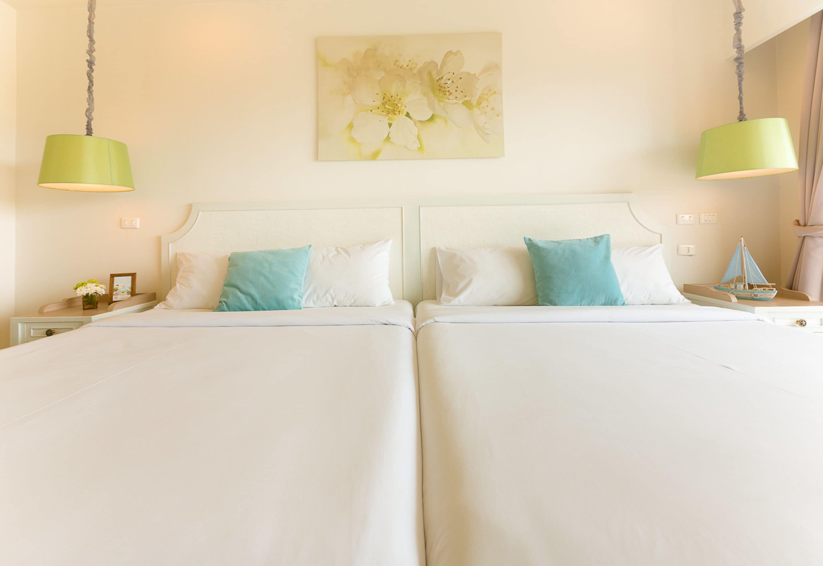A double bed with white and blue pillows, a green wall hanging, and a nightstand with a lamp.
