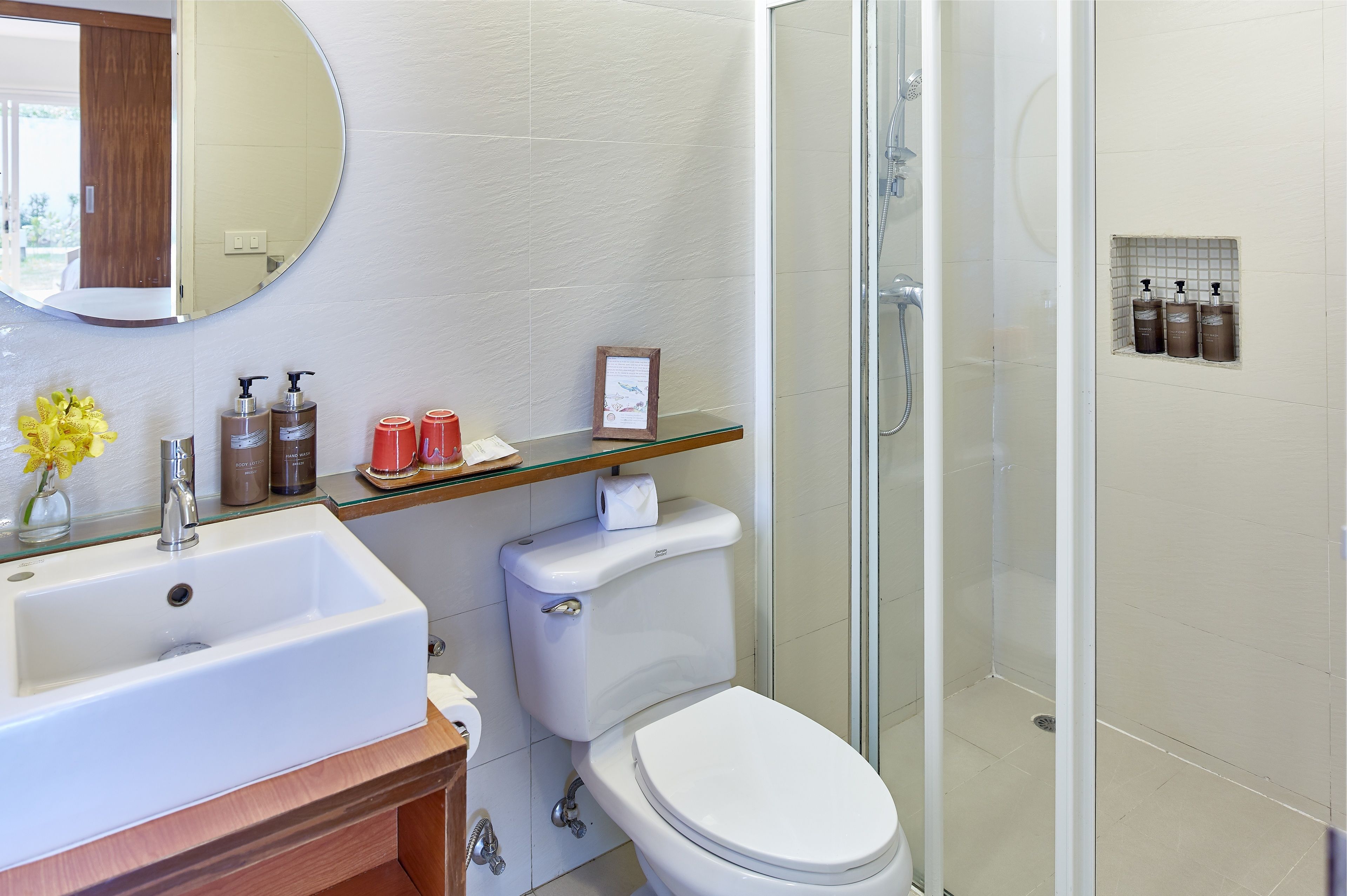 A bathroom with a white sink, toilet, and glass-enclosed shower. There are toiletries on the sink and a small shelf above the toilet.