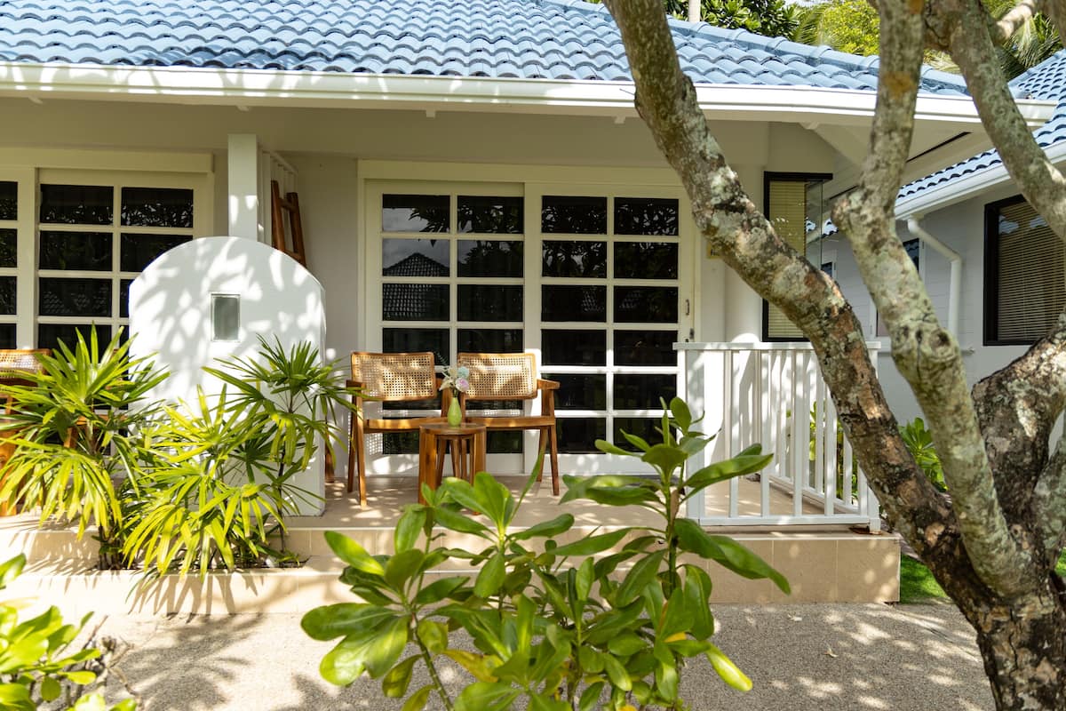 A house with a tiled roof, a porch with two wooden chairs and a table, and various potted plants.