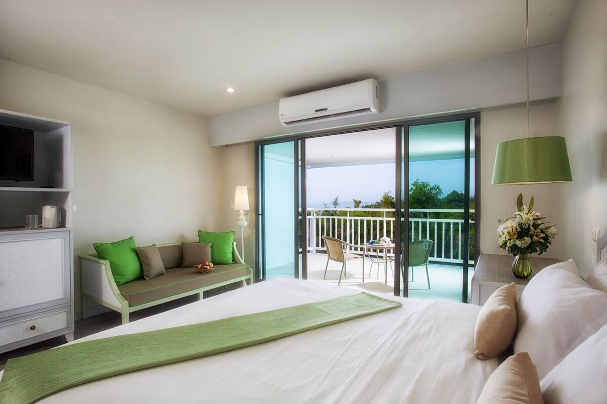 A hotel room with a bed, a sofa, a balcony with a table and chairs, and an air conditioning unit.